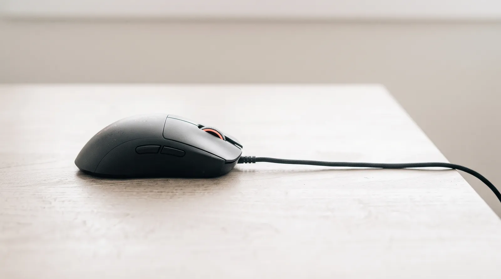 Une souris d'ordinateur filaire posée sur un bureau noir, le câble tendu vers l'ombre, sous une seule lumière corail rasante