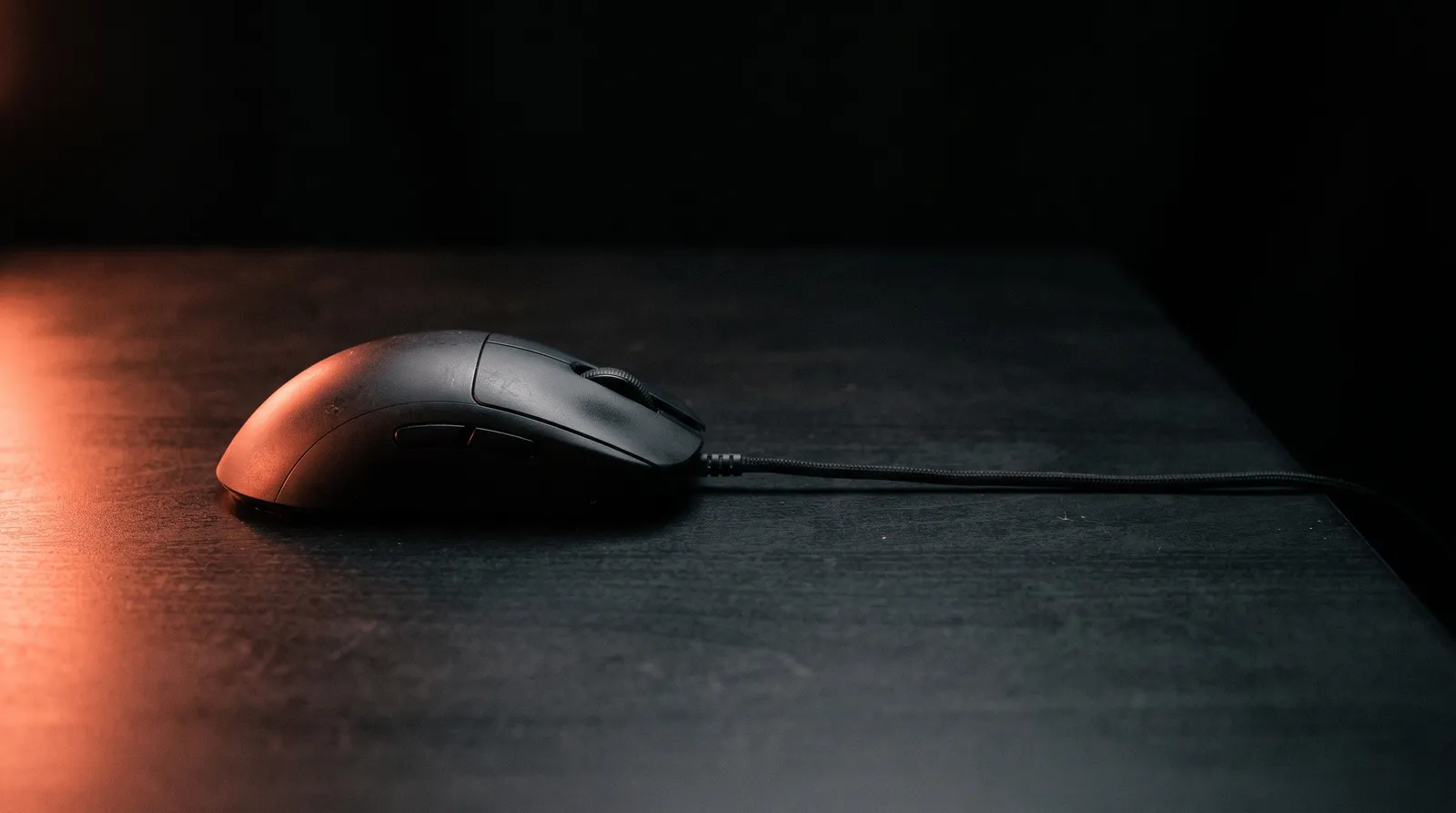 Une souris d'ordinateur filaire posée sur un bureau noir, le câble tendu vers l'ombre, sous une seule lumière corail rasante
