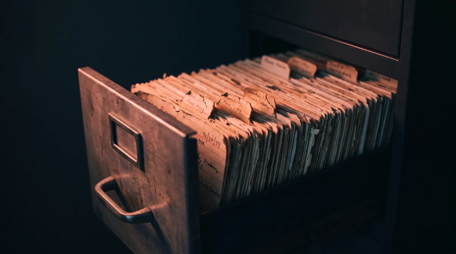 Un tiroir de classeur métallique entrouvert, rempli de fiches cartonnées, dans le noir, éclairé par une seule lumière corail rasante