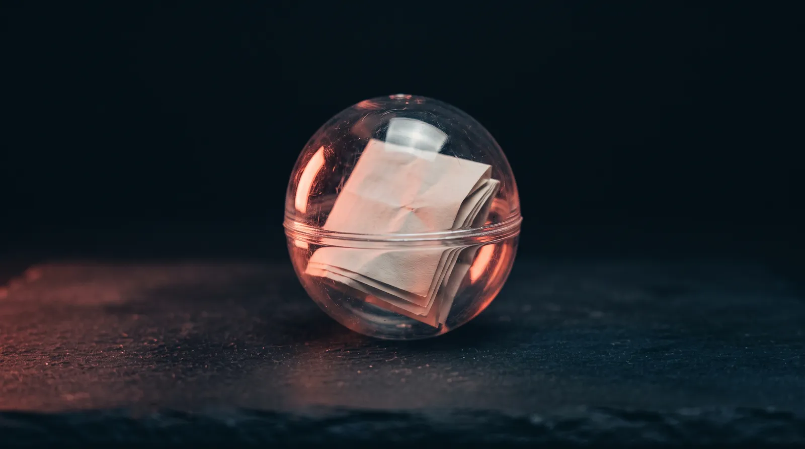 Une capsule de plastique transparent, comme celles des distributeurs, contenant une feuille de papier pliée, posée sur une table sombre et éclairée par une seule lumière corail