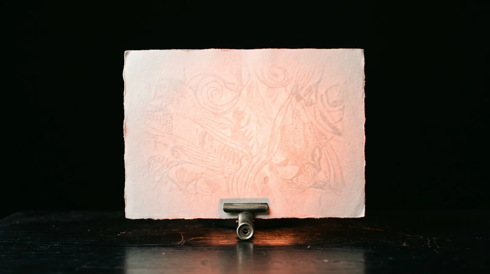 Une feuille de papier vierge dressée dans une pince métallique sur une table noire, éclairée à contre-jour par une lumière corail qui révèle un filigrane dans la fibre