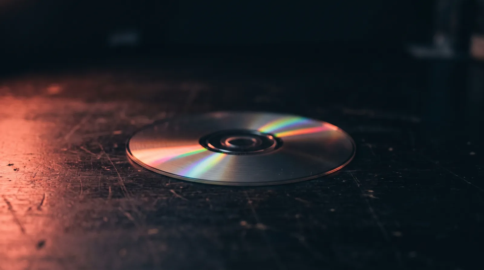 Un CD-ROM posé sur un établi sombre, sa surface irisée accrochant une seule lumière corail rasante