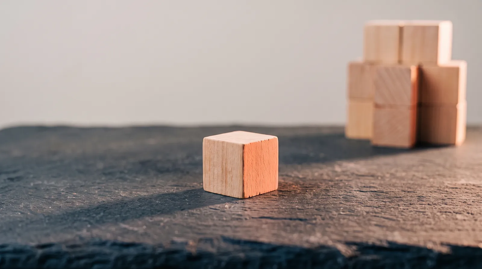 Un cube de bois isolé, poussé à l'écart d'un empilement de cubes identiques resté en arrière-plan, sur une surface d'ardoise sombre éclairée par une seule lumière corail rasante
