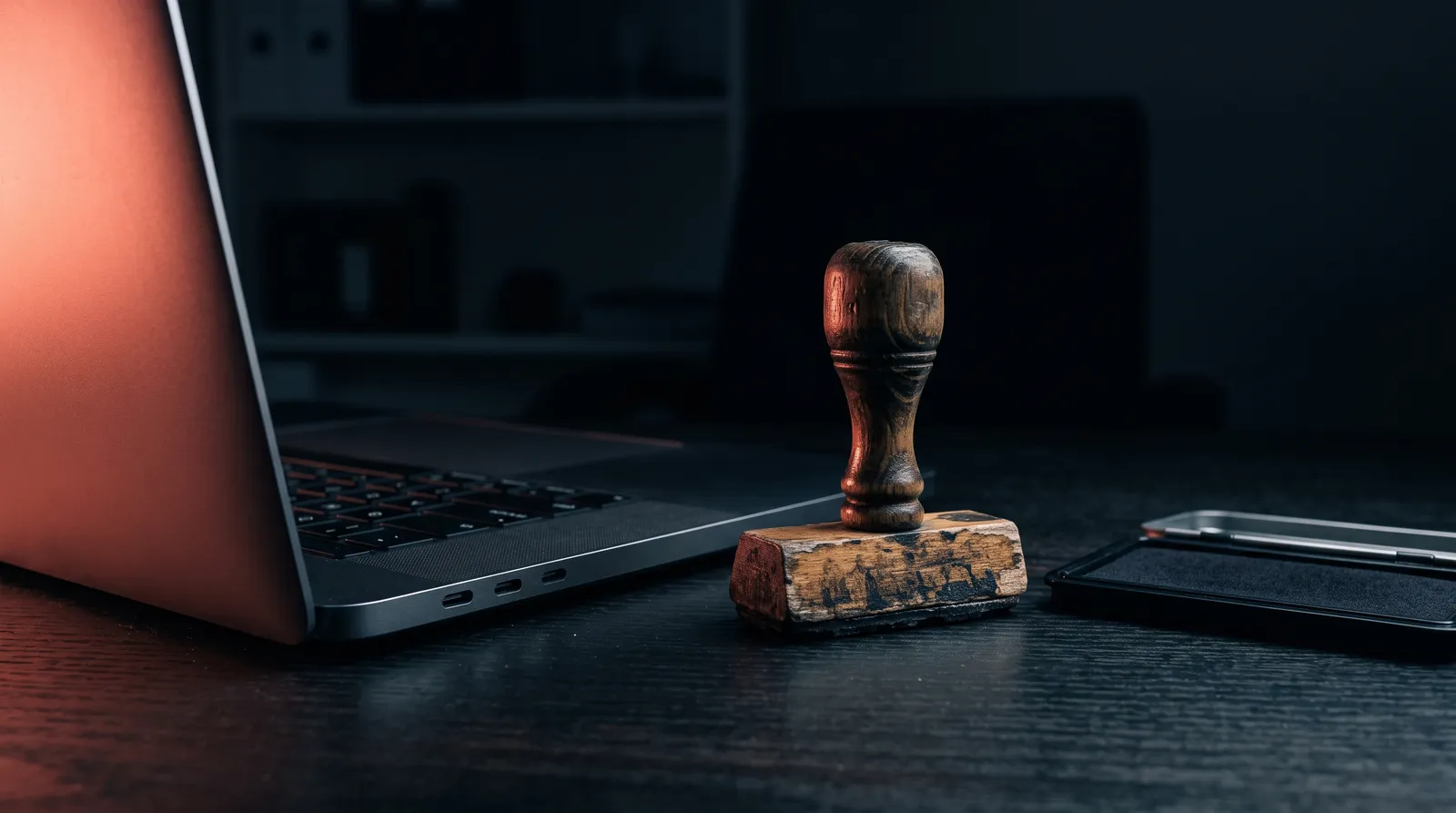 Un tampon administratif en bois posé à côté d'un clavier d'ordinateur portable, dans le noir, éclairé par une seule lumière corail