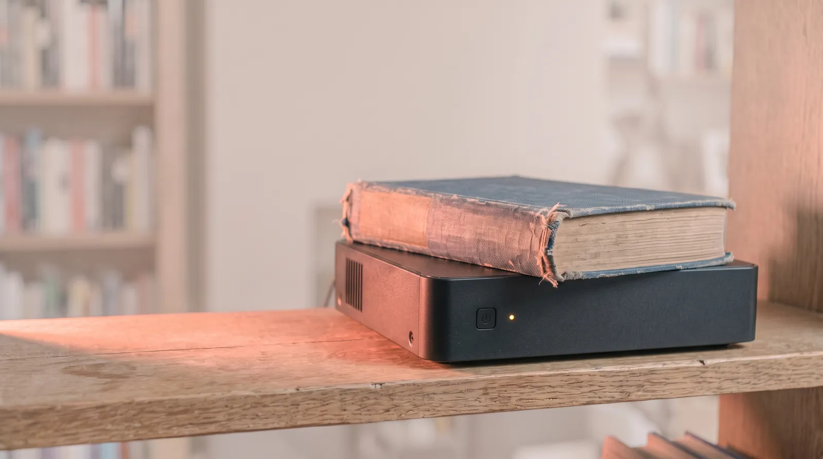 Un vieux livre relié de toile posé sur un petit boîtier serveur noir, sur une étagère en bois, éclairé par une seule lumière corail rasante