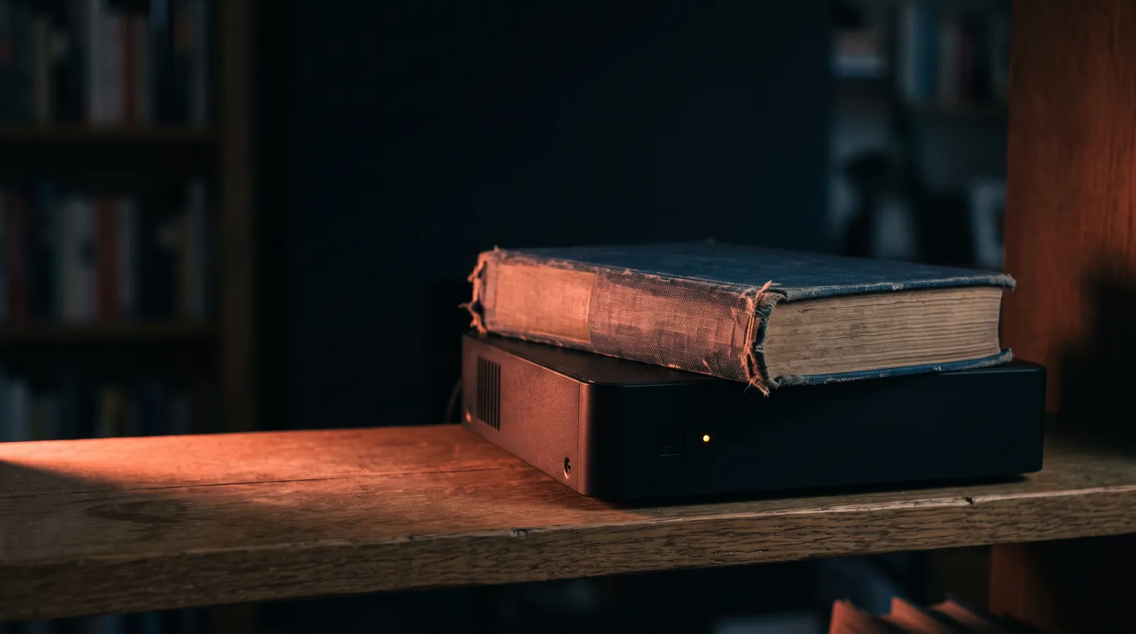Un vieux livre relié de toile posé sur un petit boîtier serveur noir, sur une étagère en bois, éclairé par une seule lumière corail rasante