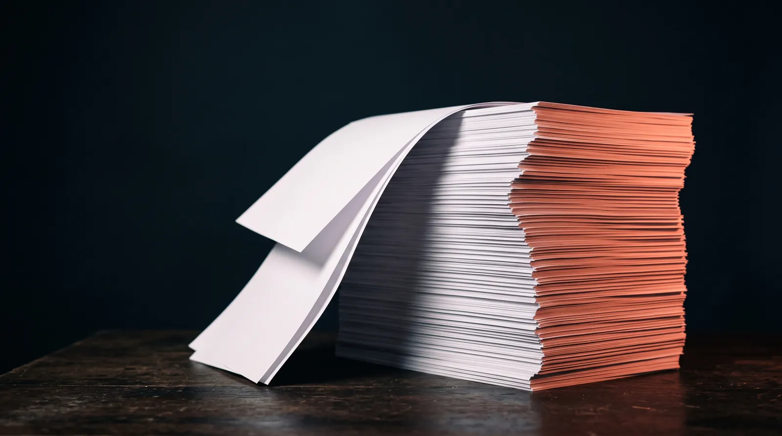 Une liasse de pages imprimées identiques, empilée dans le noir, éclairée par une seule lumière corail rasante