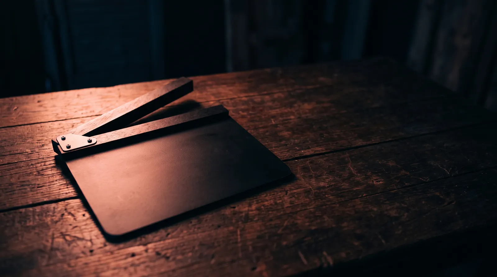 Un clap de cinéma noir posé à plat sur un bureau sombre, l'ardoise vide, éclairé par une seule lumière corail rasante