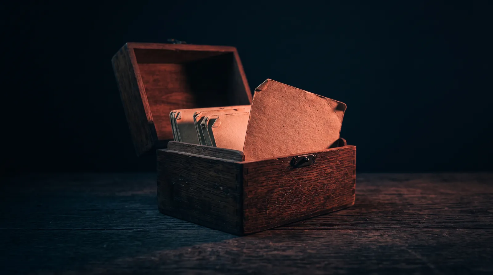 Une boîte à fiches en bois ouverte sur une table sombre, des fiches cartonnées usées dressées à l'intérieur, éclairées par une seule lumière corail rasante