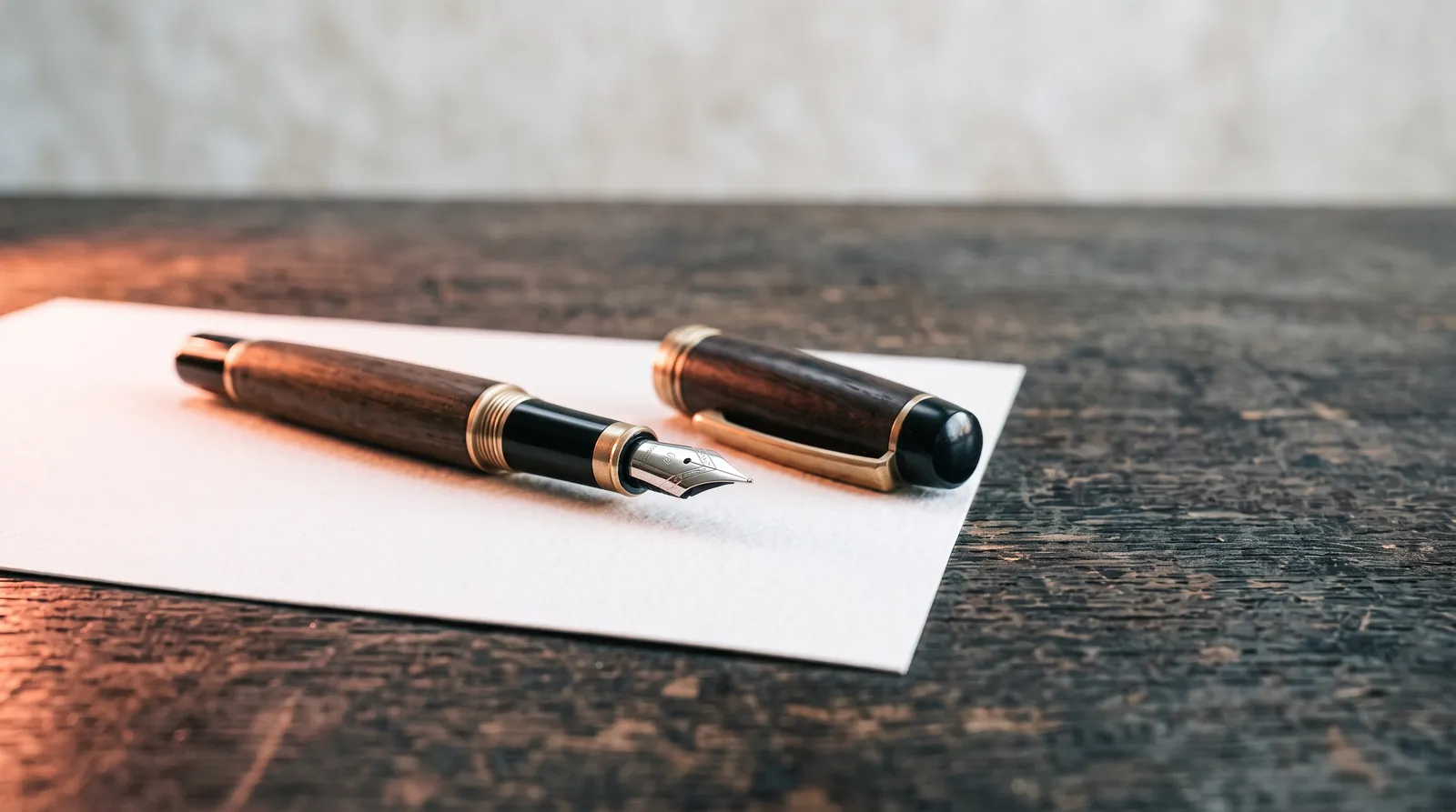 Un stylo plume ouvert, capuchon posé à côté, sur une feuille de papier blanche parfaitement vierge, sur un établi sombre éclairé par une seule lumière corail rasante