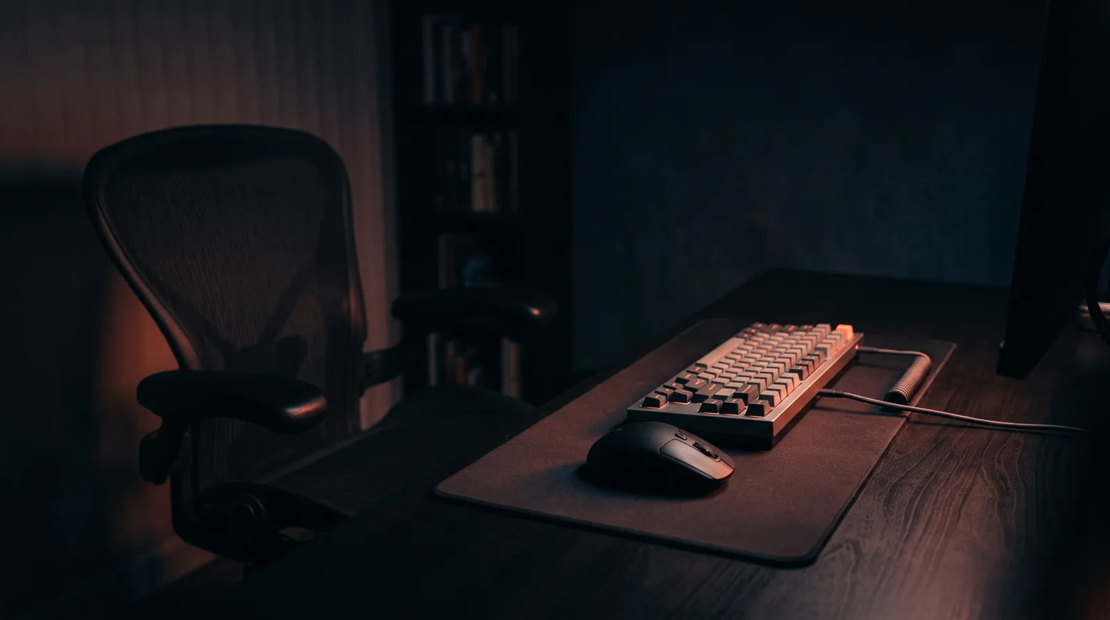 Un bureau de nuit, clavier et souris immobiles, chaise vide reculée, la lueur corail d'un écran hors champ éclairant le plateau