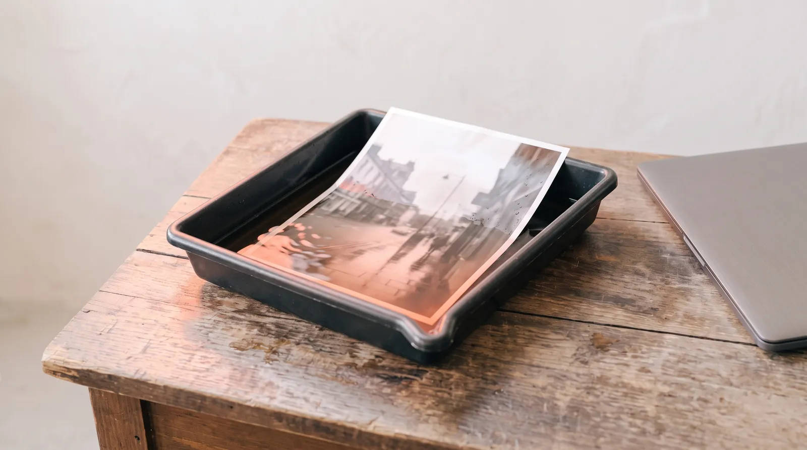Un bac de développement photo posé sur un vieux bureau en bois, un tirage qui apparaît dans le révélateur, le bord d'un ordinateur portable fermé, dans le noir, sous une seule lumière corail