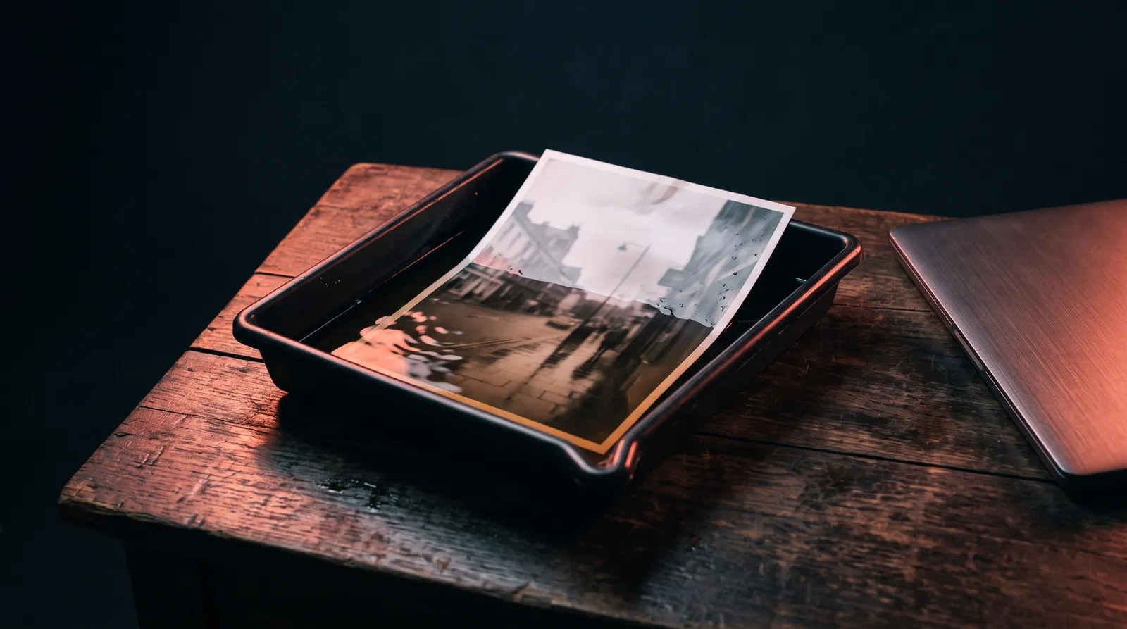 Un bac de développement photo posé sur un vieux bureau en bois, un tirage qui apparaît dans le révélateur, le bord d'un ordinateur portable fermé, dans le noir, sous une seule lumière corail