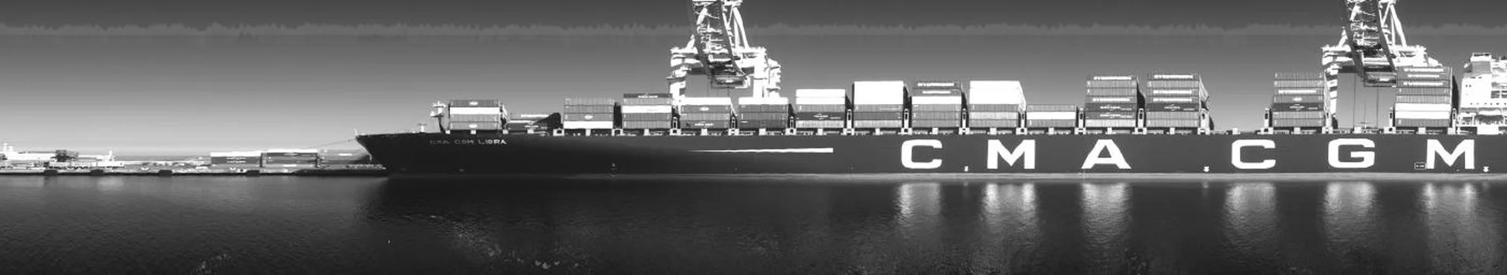 Vue en noir et blanc du port d'Oakland prise depuis un ferry : un porte-conteneurs à quai sous deux portiques, ses conteneurs empilés sur le pont, le nom de la compagnie en très grandes lettres sur la coque, et les reflets des lumières dans l'eau calme