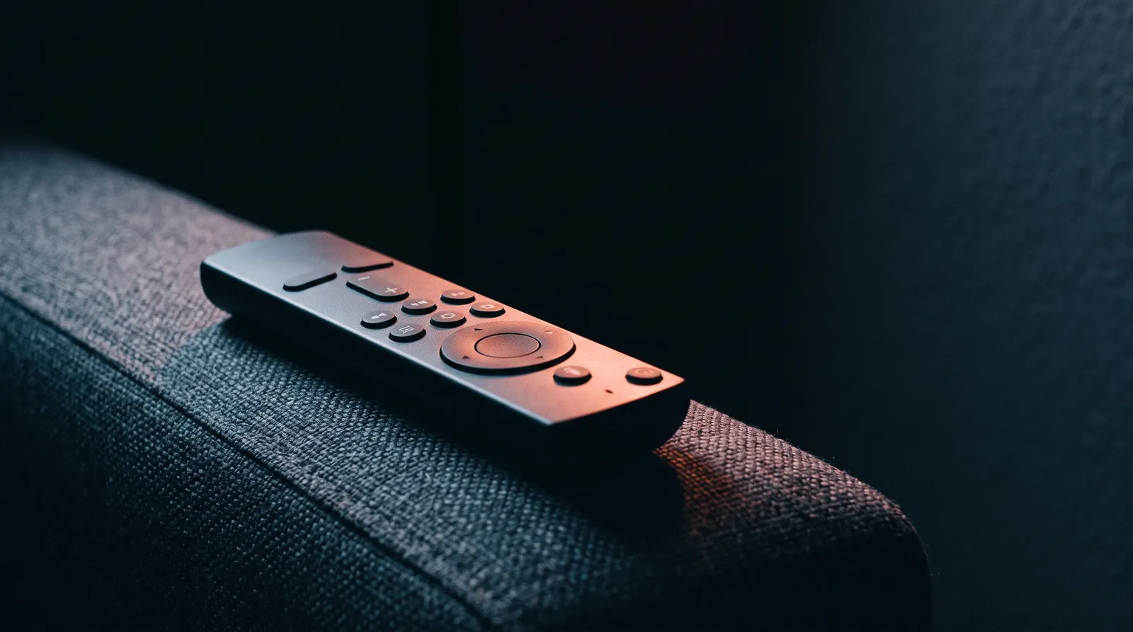 Une télécommande de télévision posée sur l'accoudoir d'un canapé dans l'obscurité, éclairée par une seule lumière corail rasante