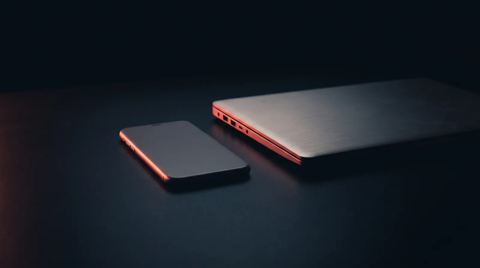 Un téléphone à l'écran éteint posé sur un bureau sombre, à côté d'un ordinateur portable fermé, aucun câble entre les deux, une seule lumière corail rasante