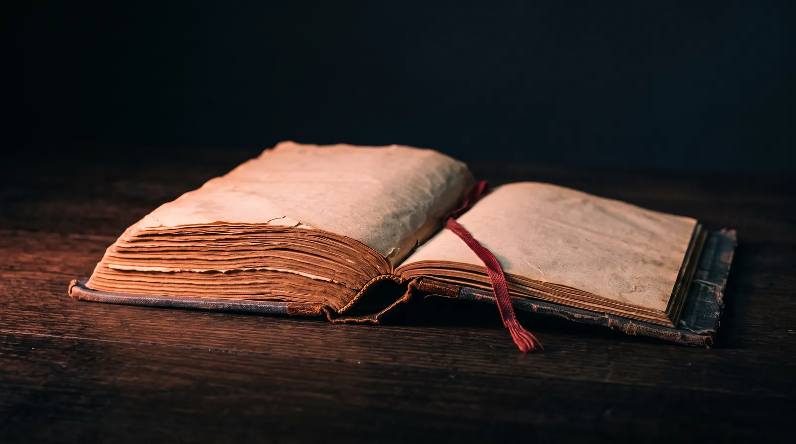 Un vieux livre relié ouvert à plat sur une table, le gros des pages lues à gauche et une poignée de pages restantes à droite, un signet en ruban sortant de la reliure, dans le noir, sous une lumière corail rasante